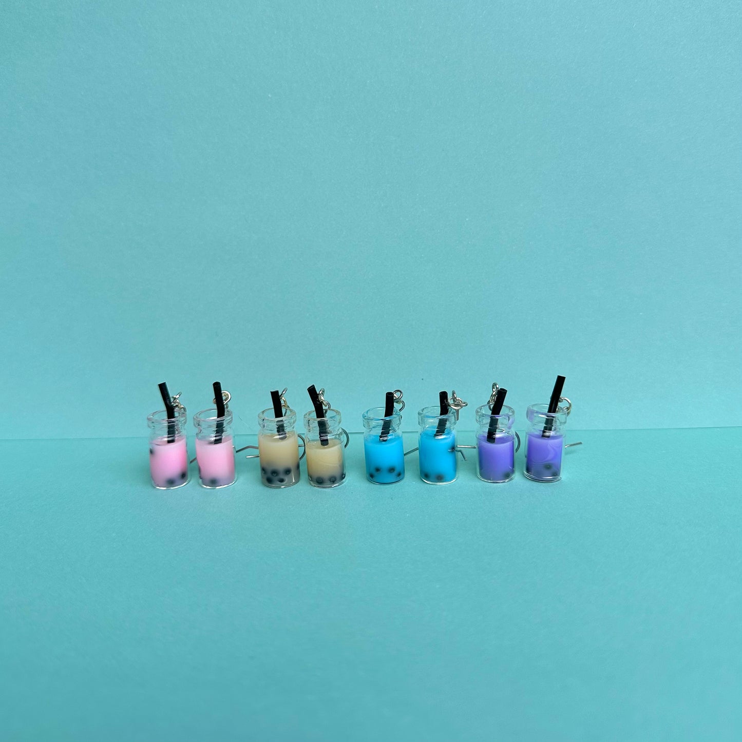 Assorted boba tea earrings in pink, beige, blue, and purple with silver hooks arranged in a row on aqua background – kawaii handmade bubble tea accessories