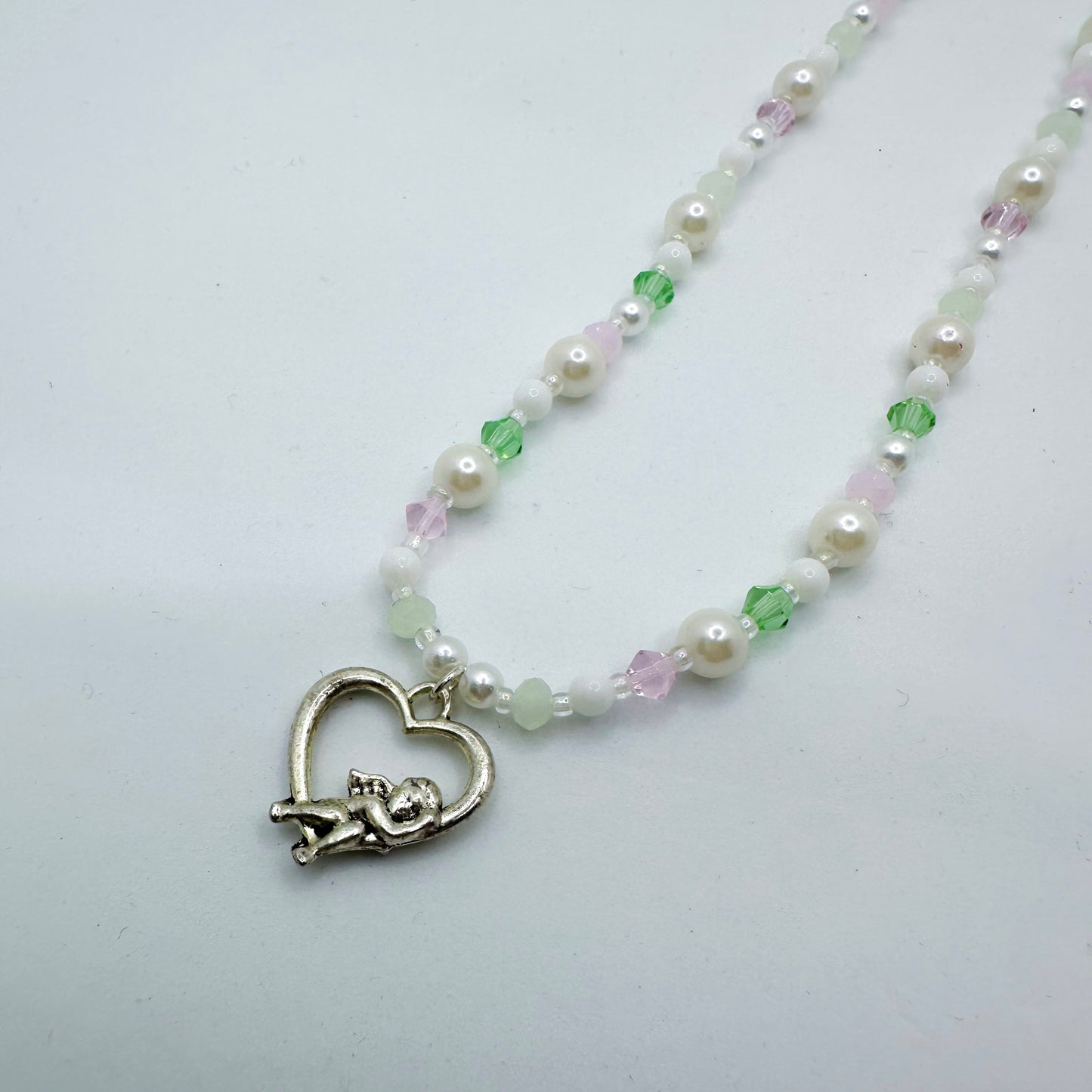 Close-up of a pastel beaded necklace with white pearls, pink and green crystals, and a silver cupid in heart charm – handmade fairycore Y2K jewellery