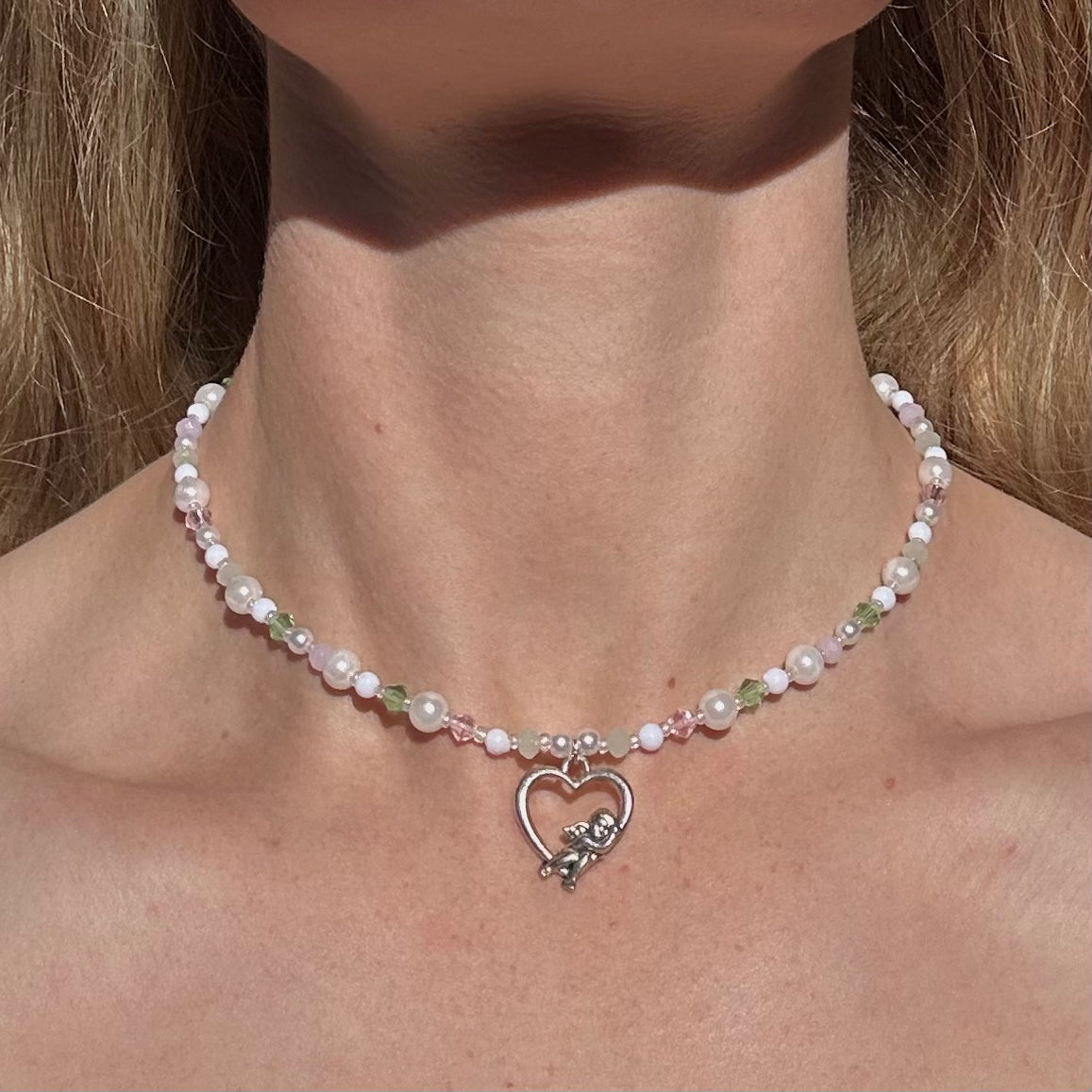 Flat lay of handmade pastel beaded necklace with pearls, green and pink crystals, and a silver cupid heart pendant – soft girl fairycore jewellery aesthetic