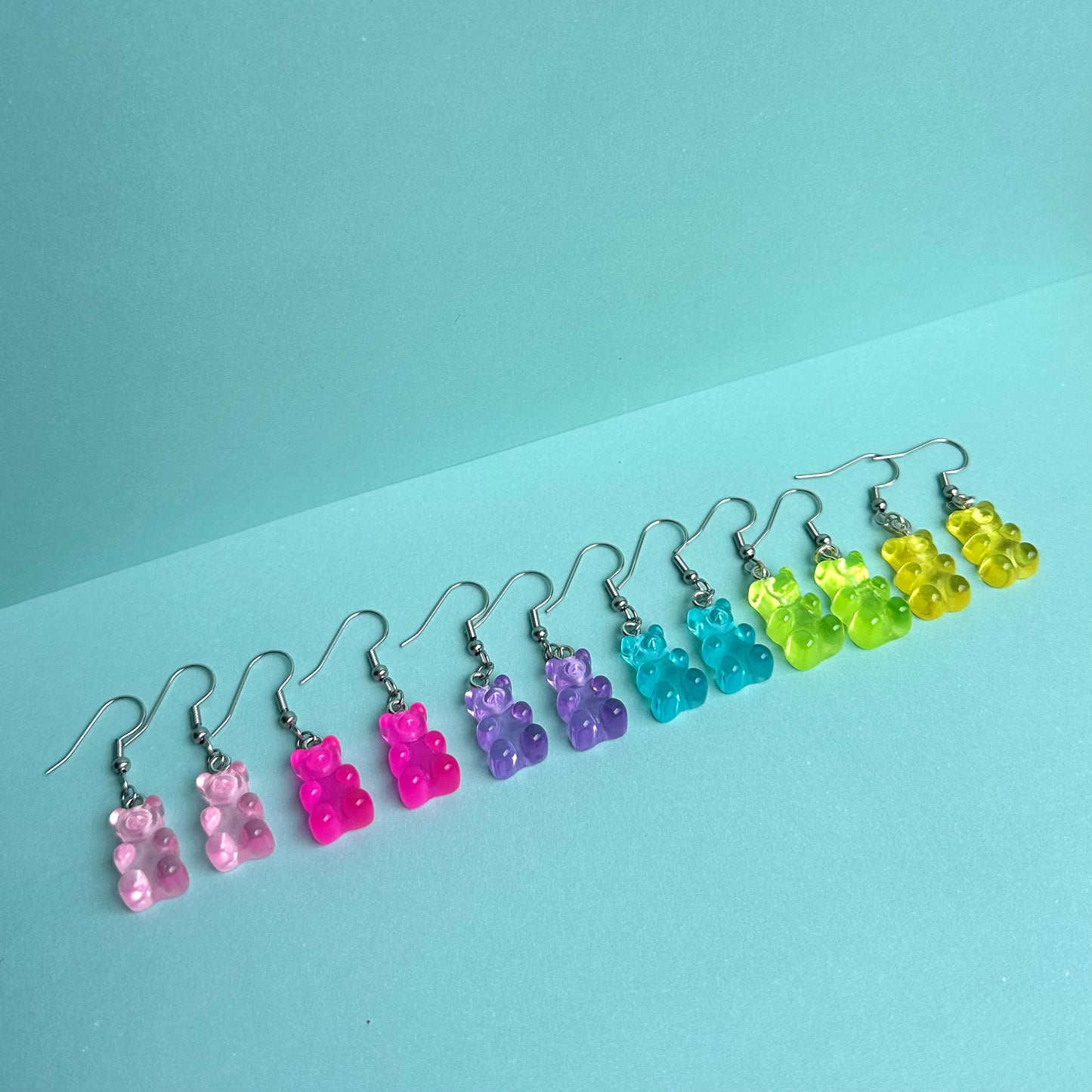 Full set of colourful gummy bear earrings in pink, purple, blue, green, and yellow arranged in a row on pastel aqua background – kawaii handmade resin jewellery by Daisy Chikk