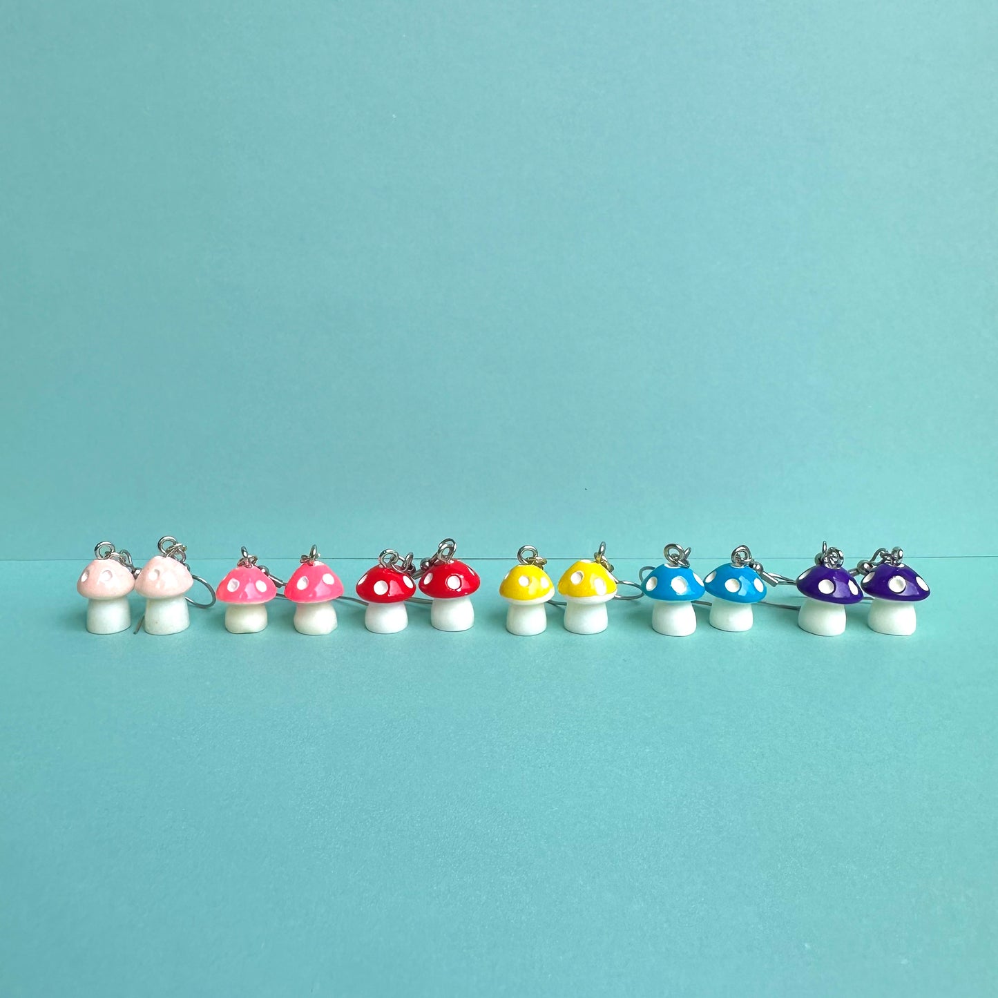 Lineup of colourful mushroom earrings in pink, hot pink, red, yellow, blue, and purple with white stems and silver hooks, displayed on aqua background – kawaii handmade jewellery