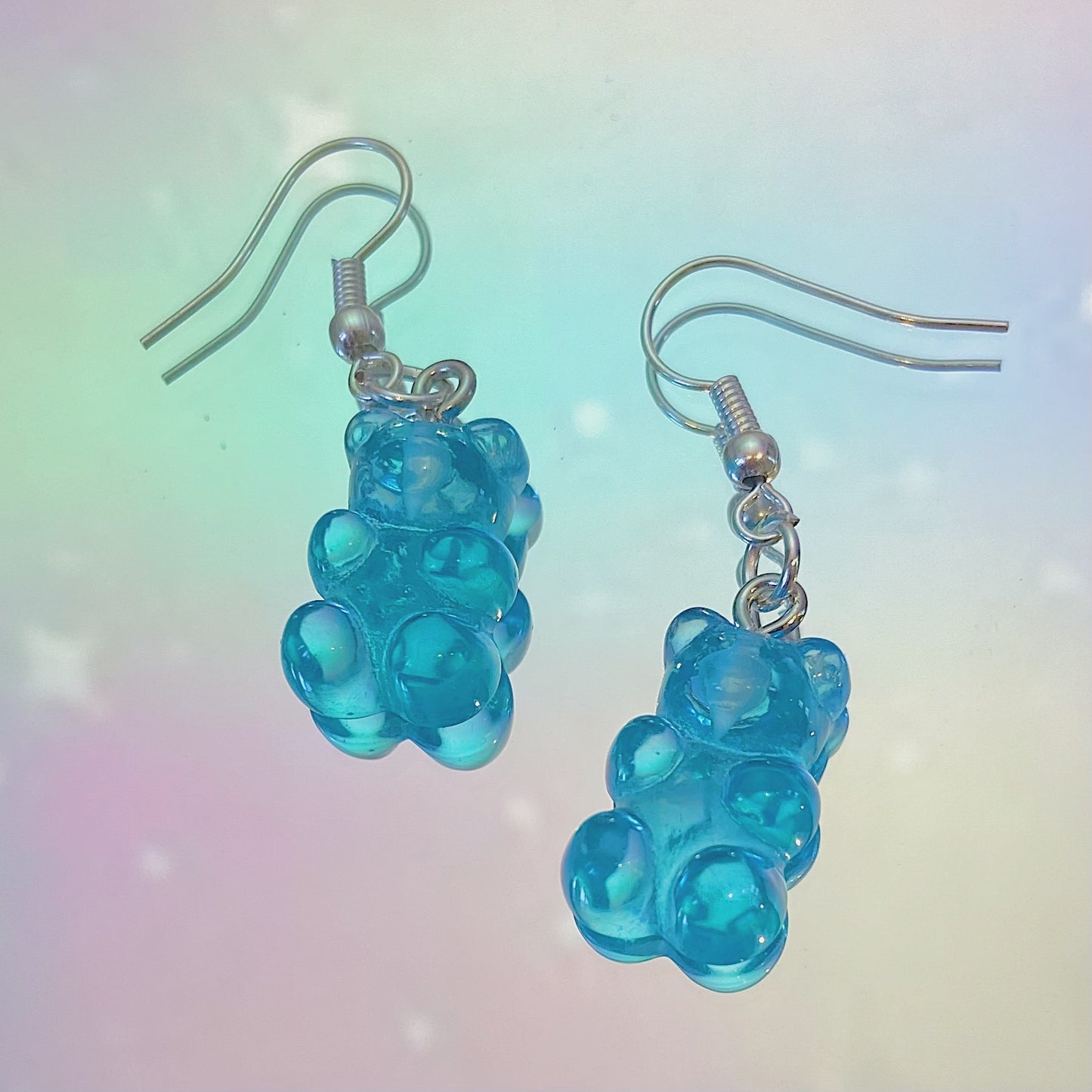 Close-up of translucent blue gummy bear earrings with silver hooks on a dreamy pastel rainbow background – kawaii Y2K handmade jewellery