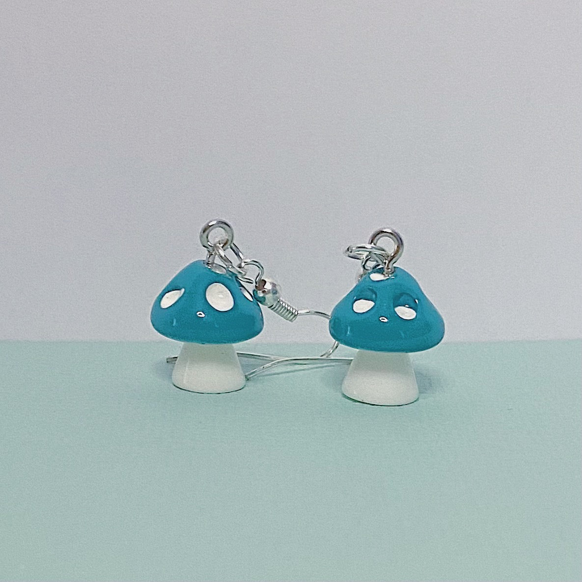 Close-up of blue mushroom earrings with white stems and silver hooks, displayed on a peach and white background – kawaii handmade toadstool jewellery