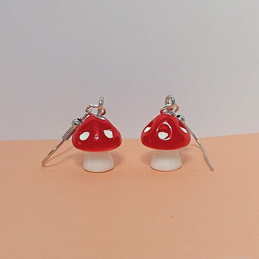 Close-up of red mushroom earrings with white stems and silver hooks, displayed on a peach and white background – kawaii handmade toadstool jewellery