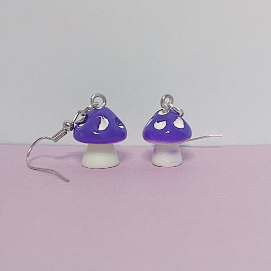 Close-up of purple mushroom earrings with white stems and silver hooks, displayed on a peach and white background – kawaii handmade toadstool jewellery
