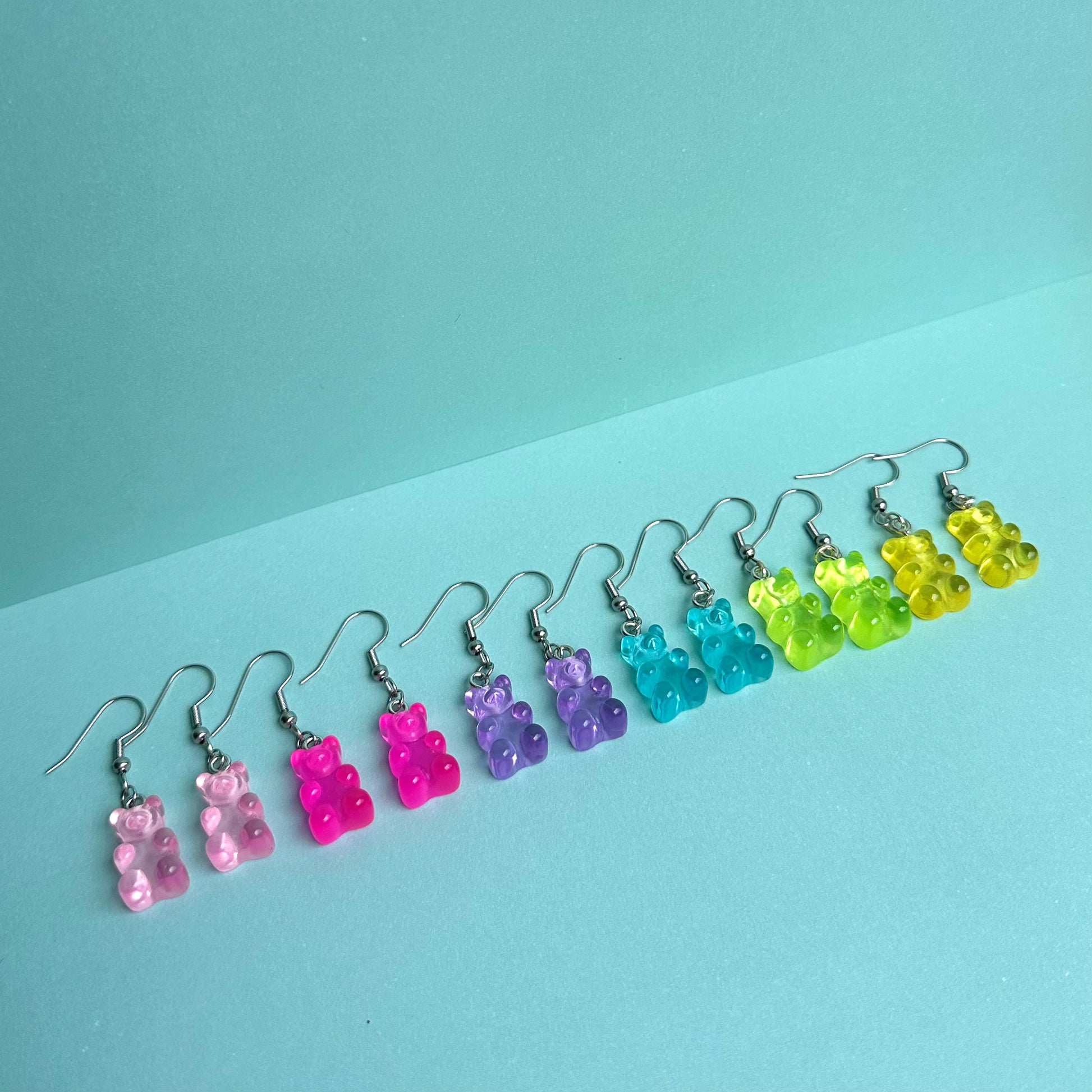 Full set of colourful gummy bear earrings in pink, purple, blue, green, and yellow arranged in a row on pastel aqua background – kawaii handmade resin jewellery by Daisy Chikk