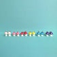 Lineup of colourful mushroom earrings in pink, hot pink, red, yellow, blue, and purple with white stems and silver hooks, displayed on aqua background – kawaii handmade jewellery