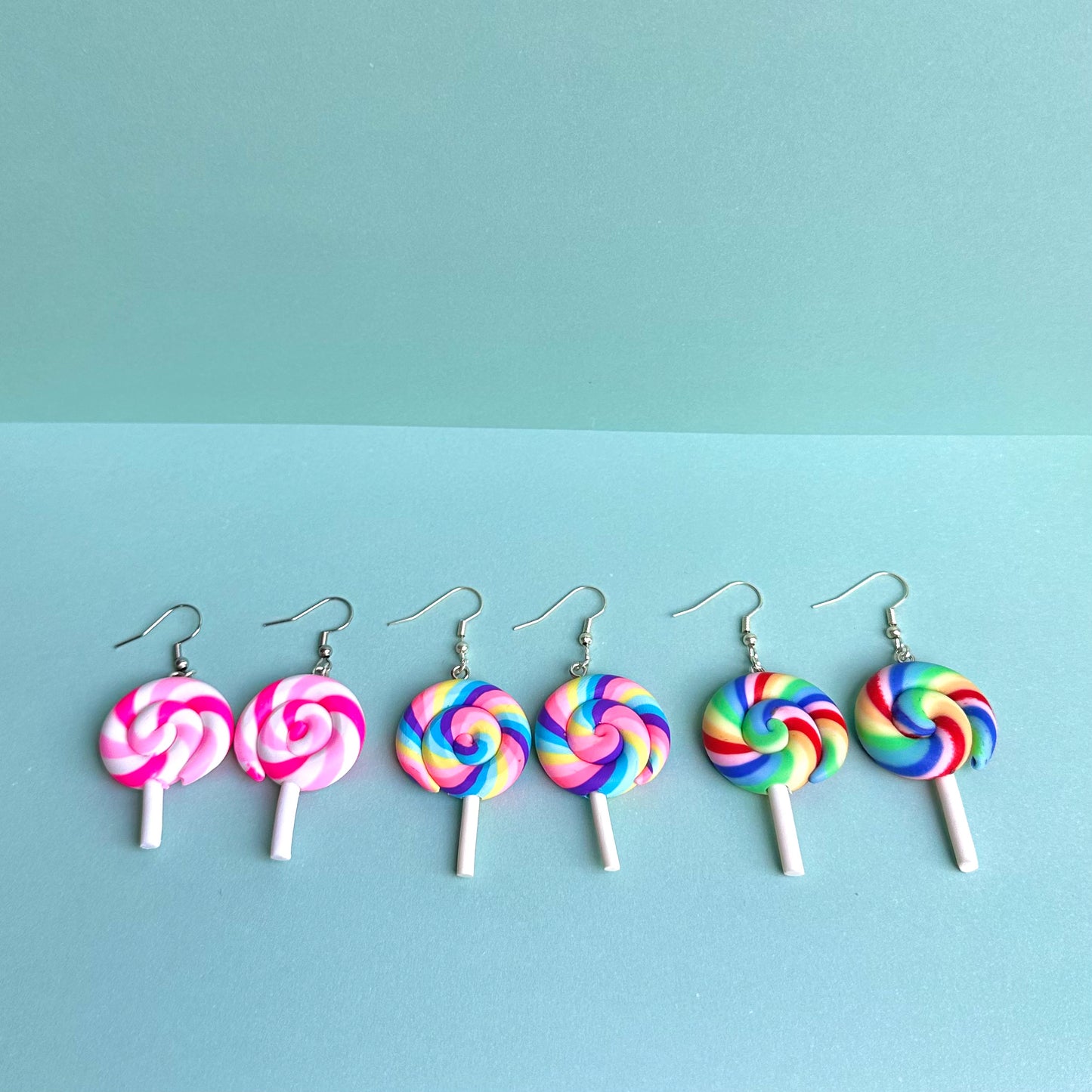 Assorted lollipop earrings in pink and rainbow swirls with white sticks and silver hooks, arranged on a pastel aqua background – kawaii handmade jewellery collection